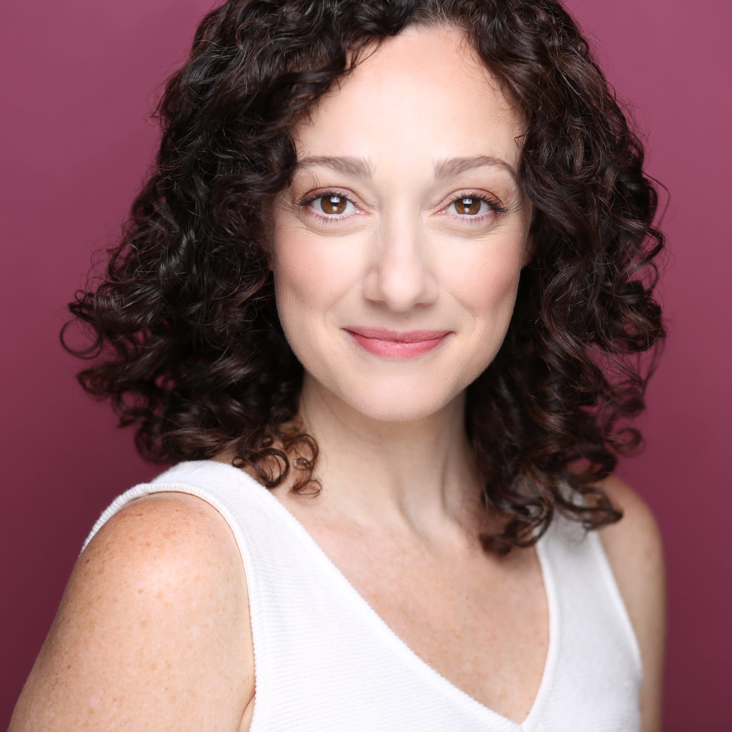 A woman with curly brown hair smiles softly against a mauve background.